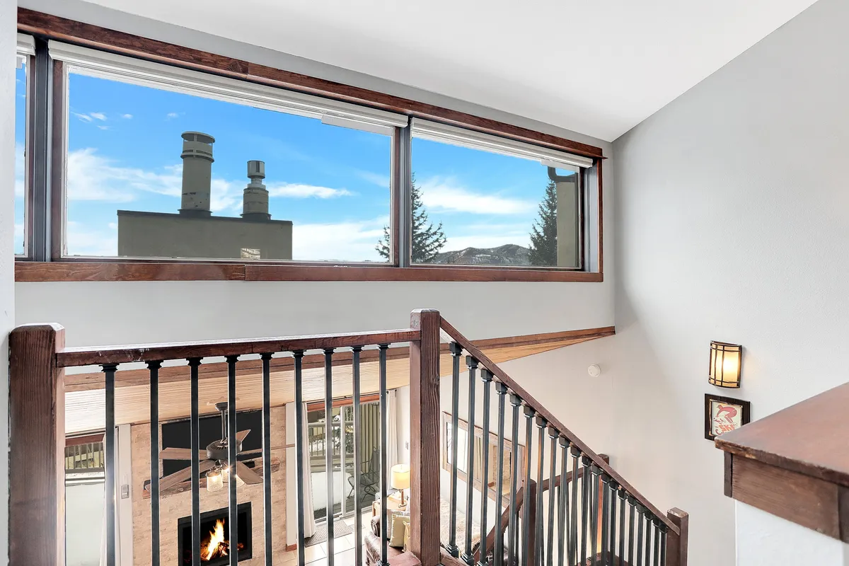 A spacious living area featuring a cozy fireplace, prominent wooden beams, and expansive windows that frame stunning mountain views. The staircase and plush seating invite relaxation and conversation, emphasizing the room's open layout and airy feel.
