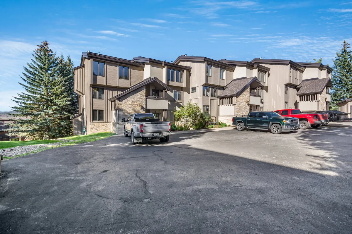 The building exterior is surrounded by tall trees and features parked vehicles, suggesting convenient access for guests with equipment. The setting feels welcoming and hints at a close-knit community atmosphere, with the mountains visible beyond, further anchoring the property in a prime location for adventure seekers.