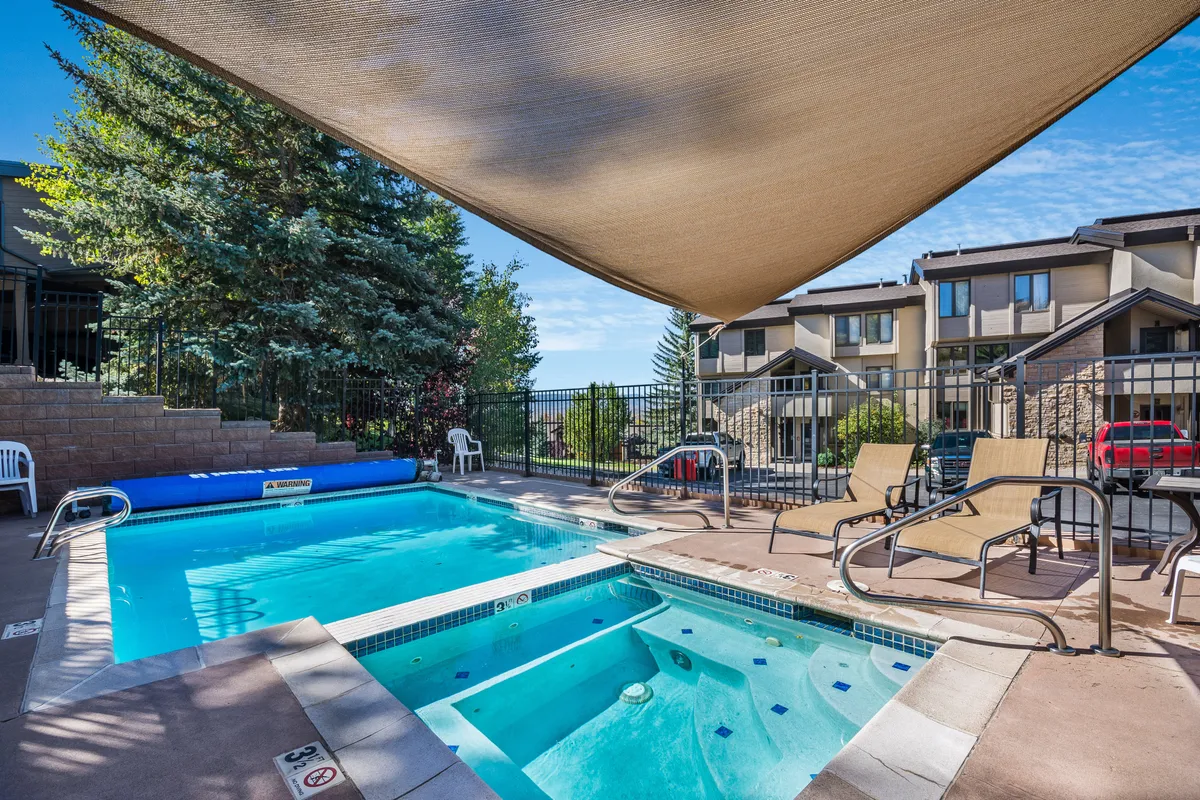 A bright, inviting pool area featuring both a swimming pool and a hot tub, surrounded by comfortable lounge chairs and a shaded area. The pool is set within a well-maintained outdoor space, perfect for socializing, swimming, and relaxation, with hints of mountain landscape in the backdrop.