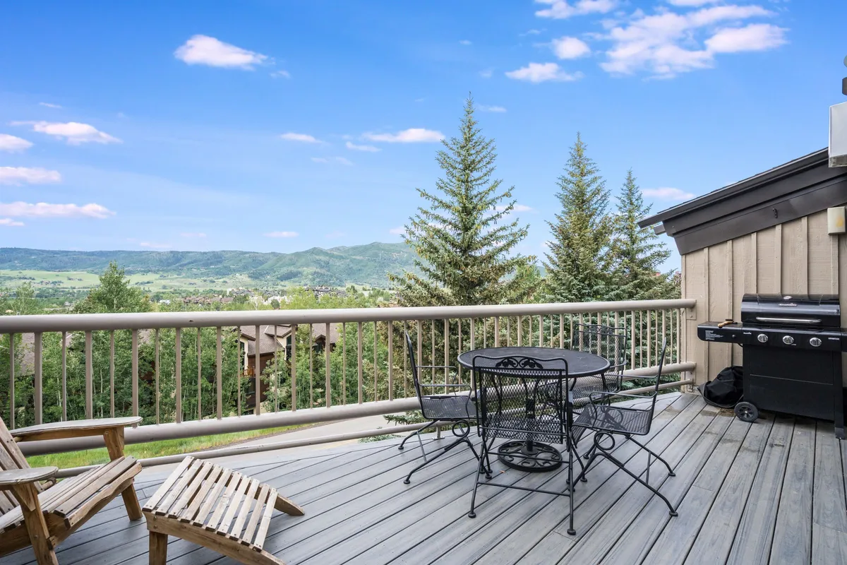 A serene balcony or deck area furnished with comfortable seating, an electric grill, and stunning mountain views framed by tall pine trees. The setup invites guests to relax, dine outdoors, or simply enjoy the scenery in any season.