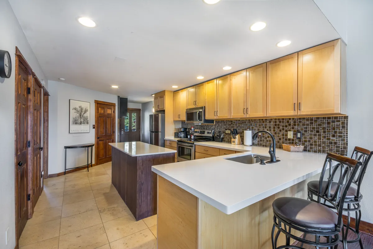 This image showcases a bright, inviting kitchen with a central island offering bar seating. The kitchen features stainless steel appliances, warm wooden cabinetry, and a striking mosaic tile backsplash. The layout is open and spacious, ideal for gatherings, casual dining, or preparing family meals together.