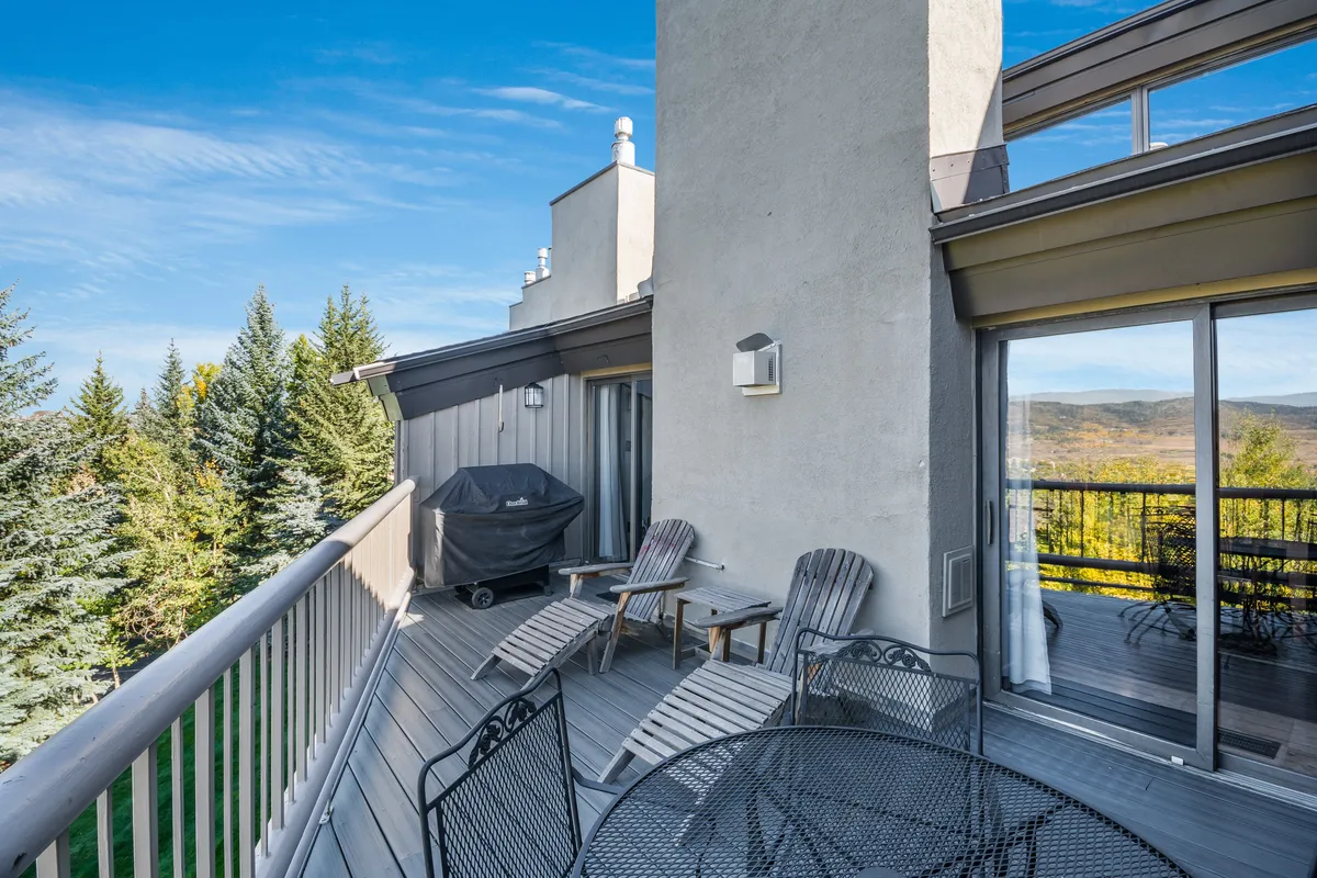 A spacious outdoor seating area featuring comfortable chairs, a grill, and a sweeping view of trees and the distant mountains. The space is well-suited for relaxing, dining, or gathering with friends after a day on the slopes. The surrounding natural landscape is tranquil and inviting, with the mountains hinting at the outdoor adventures nearby.