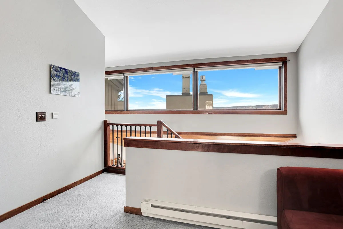A carpeted open loft area with large windows offering scenic views, a wooden railing, subtle wall art, and a dark red seating area, inviting relaxing or socializing in a semi-private nook.