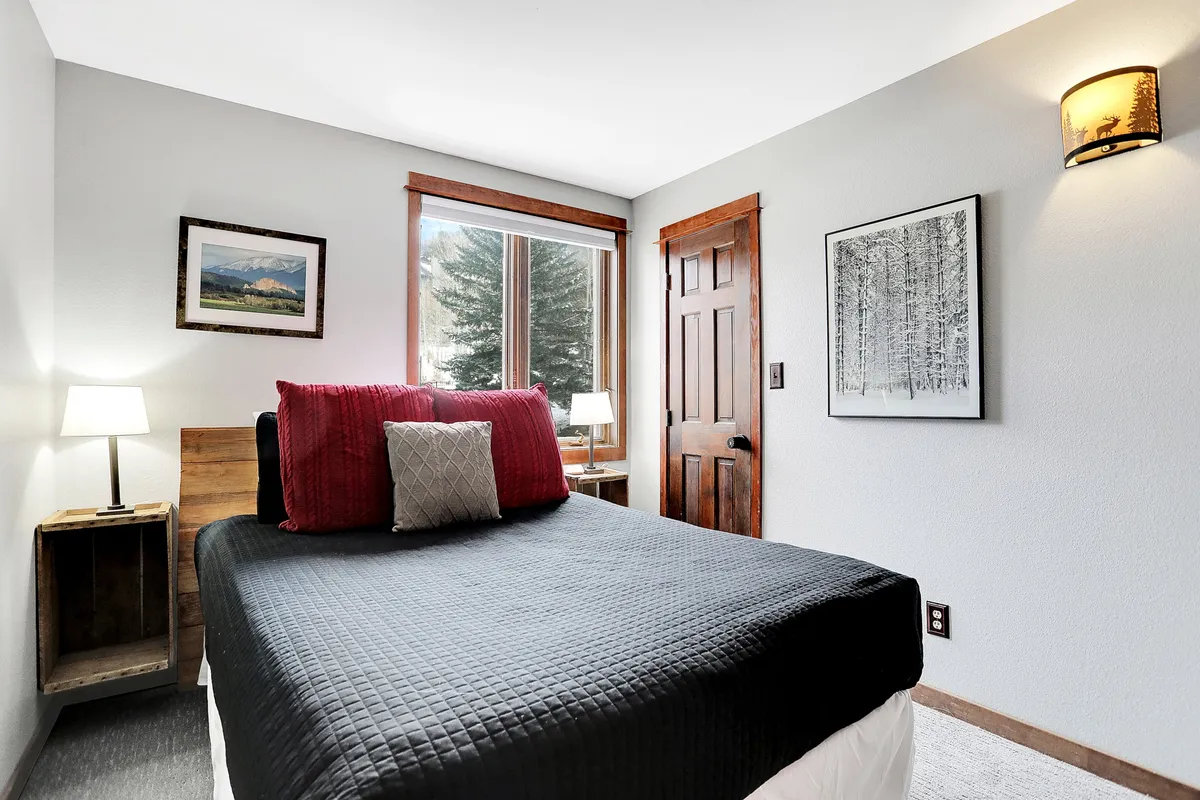 A calm, restful bedroom with a bed dressed in a black quilt and red accent pillows, a table lamp for reading, and a window offering a view outside. The setting feels intimate and designed for privacy and relaxation.