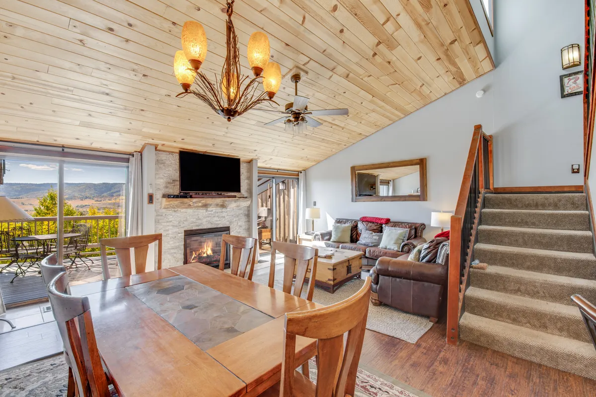 The image showcases a warm, inviting interior featuring a large dining table set for gatherings, a cozy fireplace, expansive windows framing mountain views, an elegant chandelier, and a staircase leading to the top-floor space. The soft glow of the room and the scenic backdrop create an atmosphere perfect for relaxation and quality time.