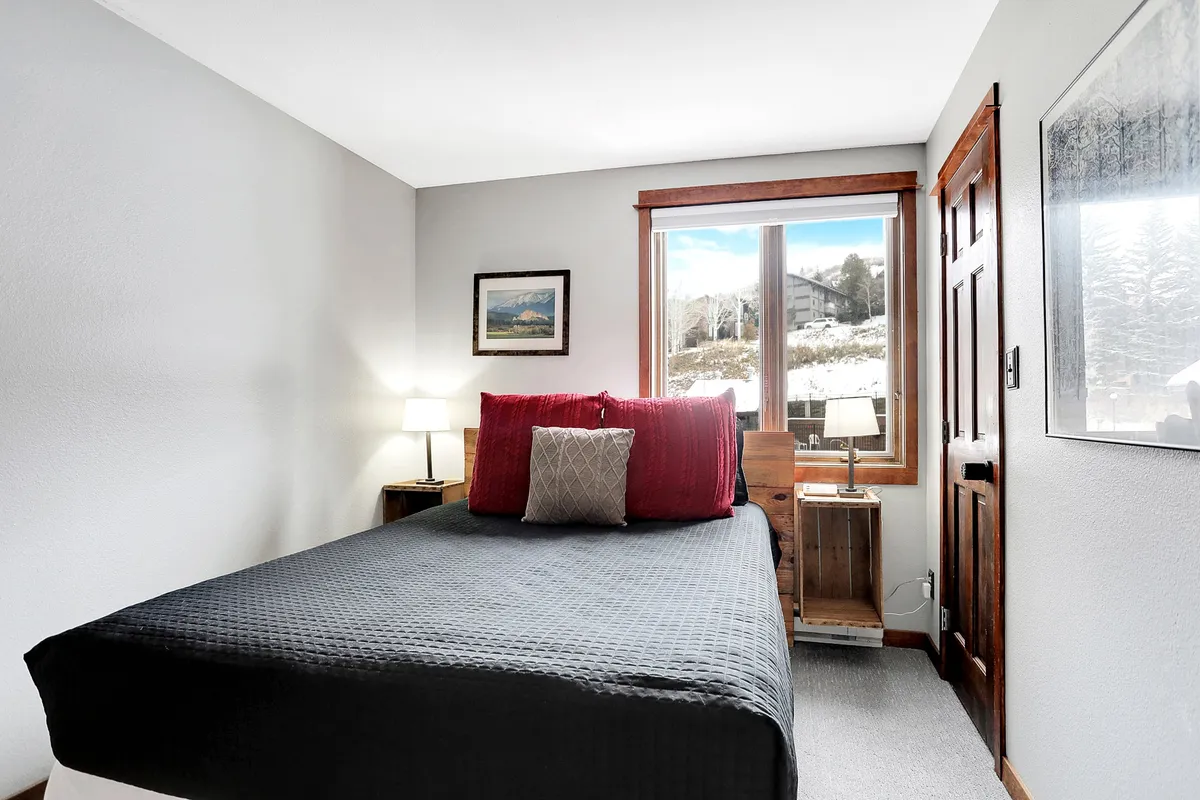This interior image features a bed with red pillows positioned by a window that offers a scenic view. The nightstand and lamp add to the room's relaxing and peaceful mood, suggesting a serene space to wake up and enjoy the surrounding scenery.