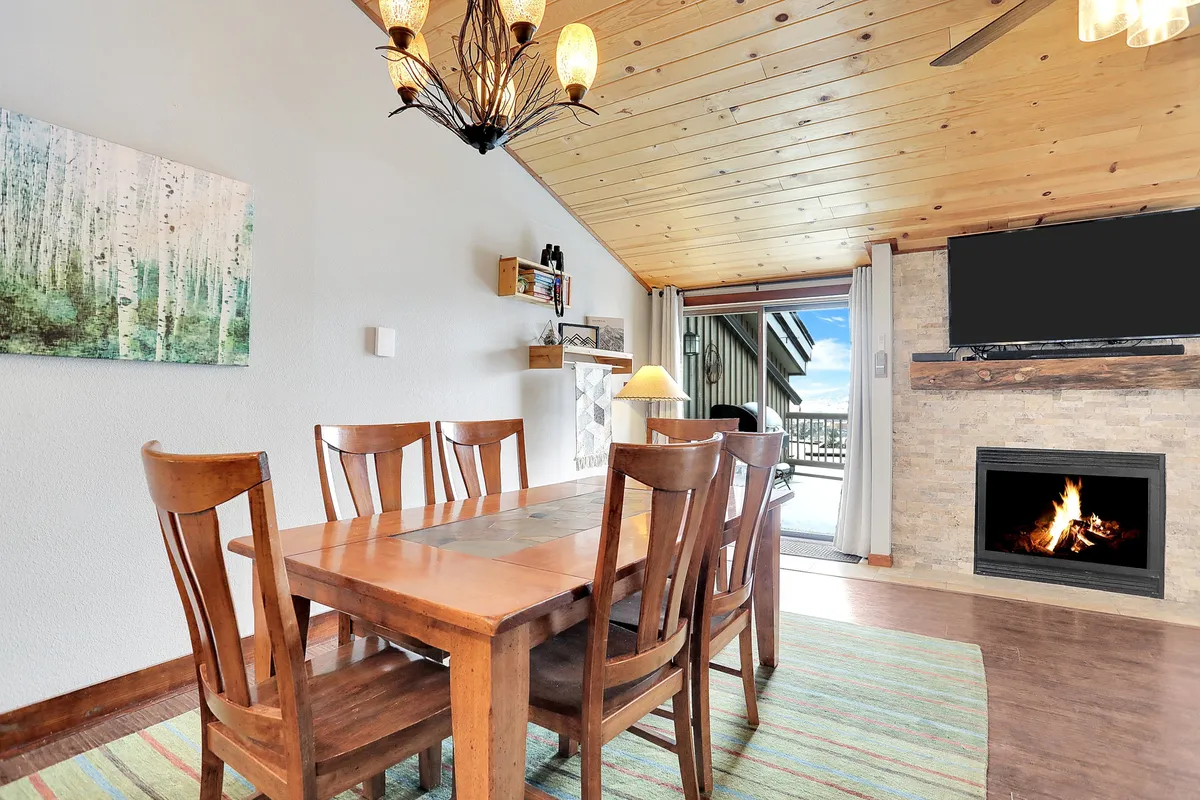 A warm, inviting interior with a dining table positioned near a fireplace and large windows. The chandelier adds a touch of rustic elegance while the windows offer glimpses of the landscape beyond, blending indoor comfort with outdoor beauty.