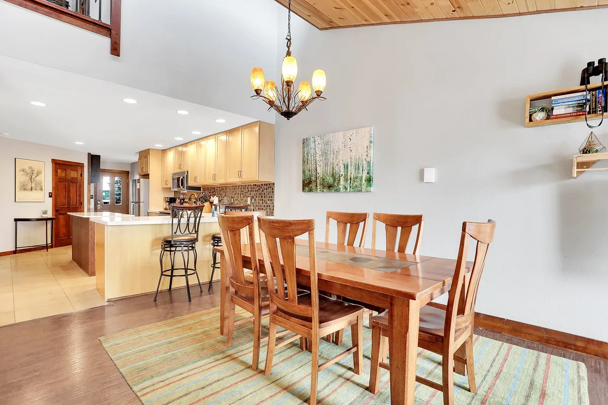 This interior shot features a well-lit dining area with a wooden table set for six, a stylish chandelier above, and modern artwork on the wall. The kitchen area is visible in the background, reinforcing the connection between dining and cooking spaces. The mood is warm and welcoming, ideal for family feasts or special celebrations.