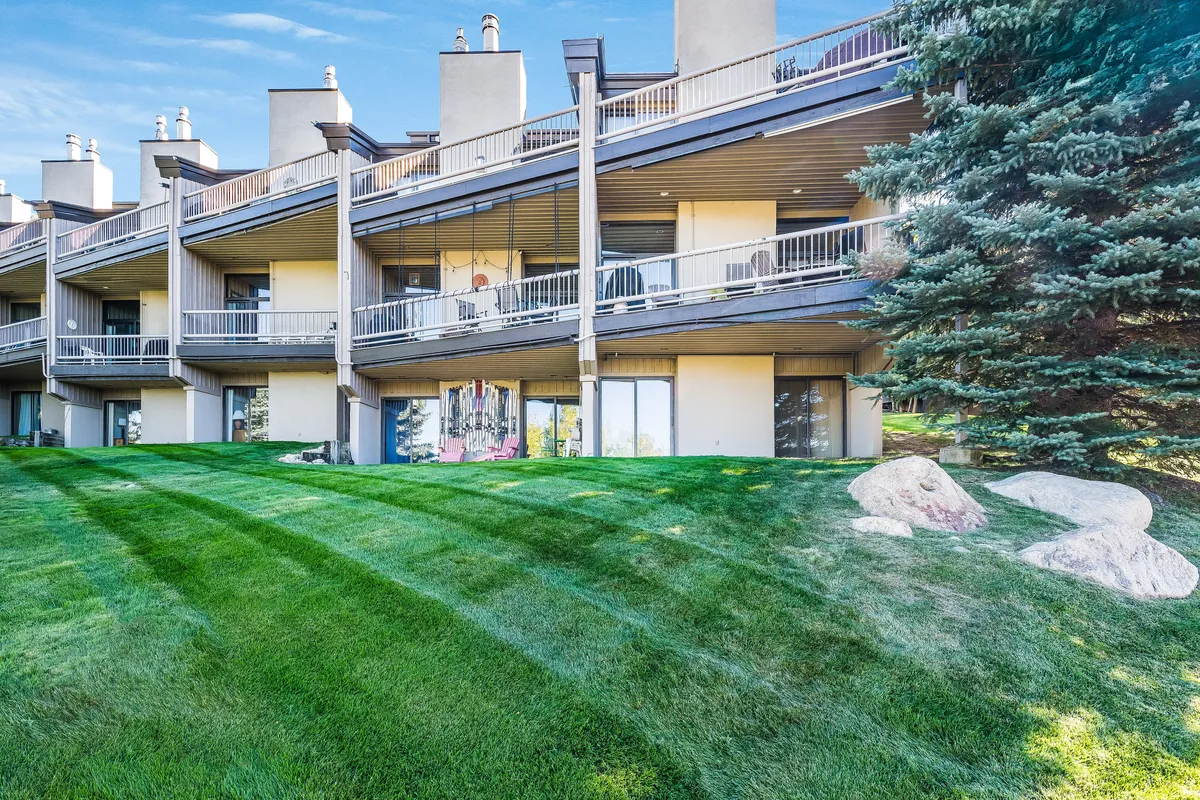 The exterior of a two-story building with multiple balconies, set amid a lush green lawn, rock outcroppings, and tall trees. The inviting setting is enhanced by peaceful, natural surroundings, evoking the mountain retreat atmosphere central to the experience.